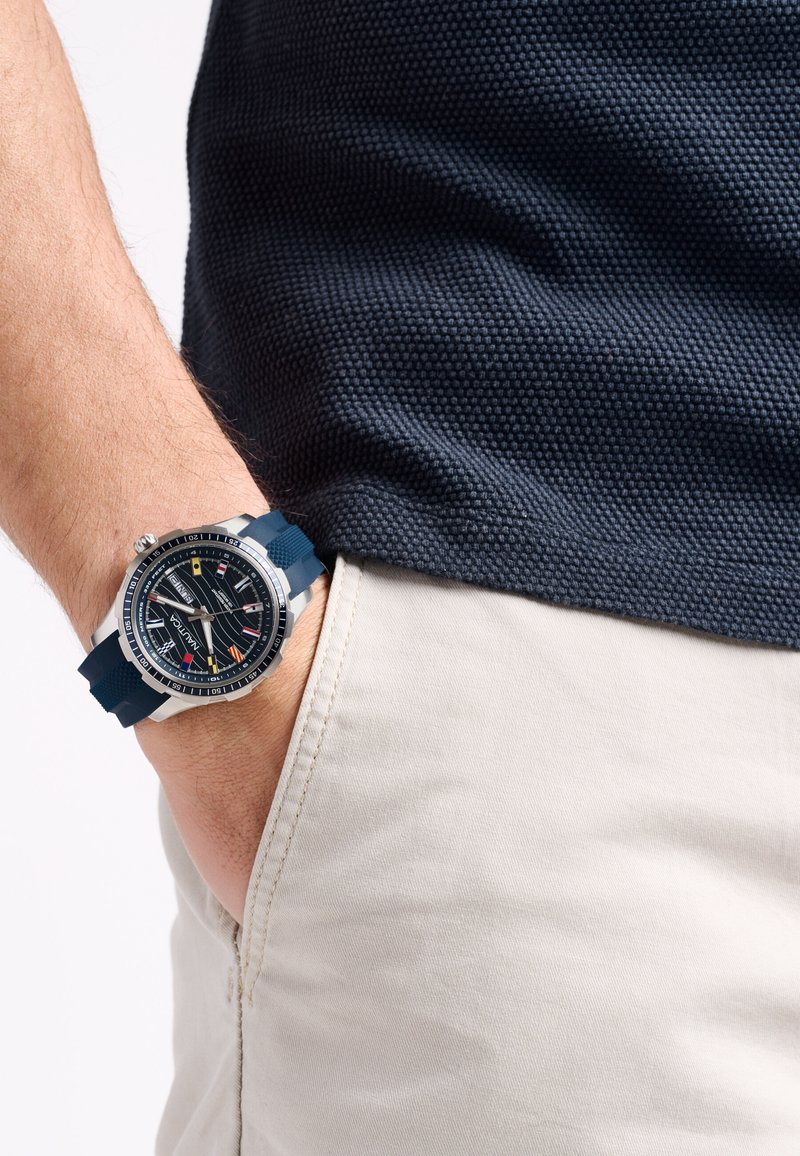 Orologio da polso nero e argento con cinturino in silicone blu navy, quadrante testurizzato, accenti colorati, esposto su una mano che riposa in una tasca beige chiaro.
