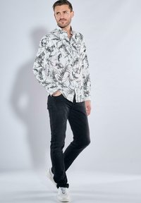 Weißes Hemd mit schwarzen Blumenmustern, lange Ärmel, Knopfleiste; kombiniert mit schwarzen Jeans und weißen Sneakers.