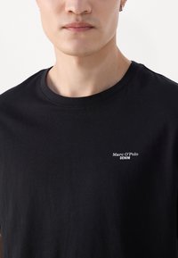 Zwarte katoenen t-shirt met een ronde hals en korte mouwen. Heeft een wit logo "Marc O'Polo DENIM" op de linkerkant van de borst.