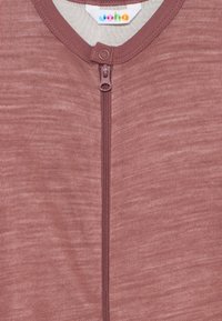 Detailný záber na fialovú zipsovú spodnú časť s okrúhlym výstrihom, viditeľný zips, cvokový gombík a farebný logotype na vnútornom štítku.