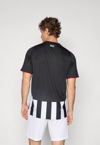 Must spordisärk punaste detailidega, lühikeste varrukatega ja 1903 graafikaga seljal. Alumises servas on triibuline muster.