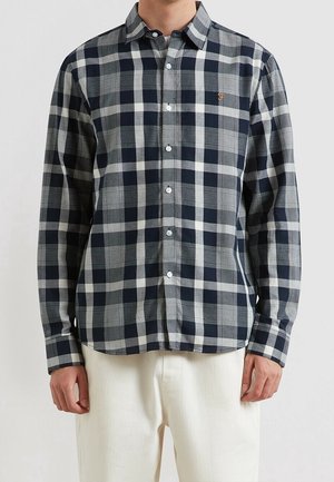 Mann trägt ein langärmeliges, schwarz-weiß-grau kariertes Hemd mit weißen Hosen und steht vor einem einfarbigen Hintergrund.