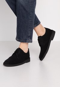 Black suede lace-up shoes with a low rubber sole, rounded toe, and stitched detailing, worn with cuffed dark denim jeans.