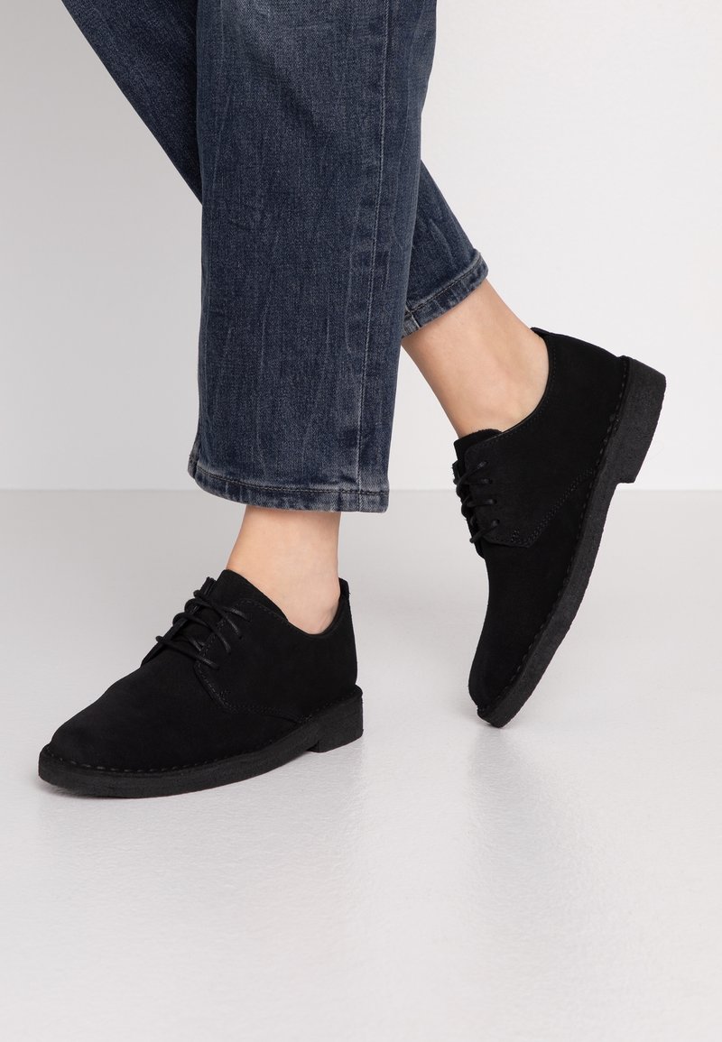 Black suede lace-up shoes with a low rubber sole, rounded toe, and stitched detailing, worn with cuffed dark denim jeans.