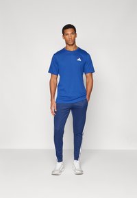 Blue athletic t-shirt with short sleeves and white logo, paired with matching blue tapered pants featuring white stripes on the sides.