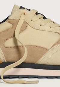 Sneaker beige e tan con materiali misti in suede e tessuto, punta arrotondata, lacci piatti beige e un accento nero sul tallone.