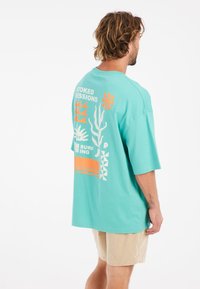 Tyrkysové oversized tričko s grafickým designem v oranžových a bílých prvcích, včetně textu, květin a abstraktních tvarů.