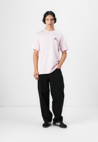 Helles rosa T-Shirt mit kurzen Ärmeln und kleinem roten Logo, kombiniert mit lockeren schwarzen Hosen und schwarzen Sneakers mit weißen Streifen.