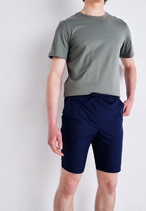 Mężczyzna w oliwkowozielonym t-shircie z krótkim rękawem i granatowych szortach ze sznurkiem stoi na tle białej, gładkiej ściany.