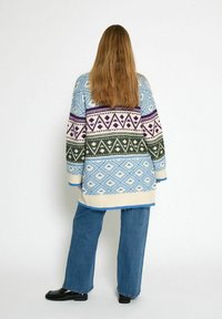 Maglione colorato lavorato a maglia con un motivo geometrico in blu, viola e verde, abbinato a jeans a gamba larga blu e scarpe nere.