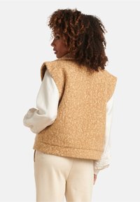 Tan wool vest featuring a textured finish, sleeveless design, and a rounded collar, worn over a white long-sleeve shirt.