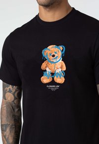 Black cotton t-shirt featuring a graphic of a teddy bear with orange fur and blue accents. Text below reads "CLOSURE LDN" and "MILANO."