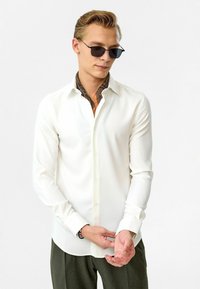 Witte lange mouwen shirt met knoopkleur, met een gemusterde binnenvoering. Model draagt een donker zonnebril en groene broek.
