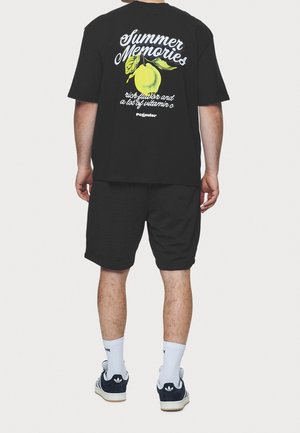 Schwarzes T-Shirt mit Grafik "Sommererinnerungen" und Zitronendesign, kombiniert mit schwarzen Shorts. Weiße Socken und marineblaue Schuhe vervollständigen das Outfit.