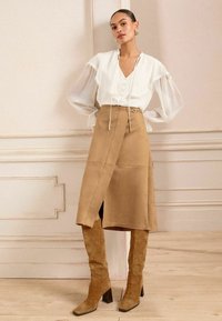 Blusa bianca con maniche trasparenti e scollatura a V, abbinata a una gonna in camoscio tan al ginocchio con un'apertura laterale e un dettaglio in cintura. Stivali alti in camoscio tan.