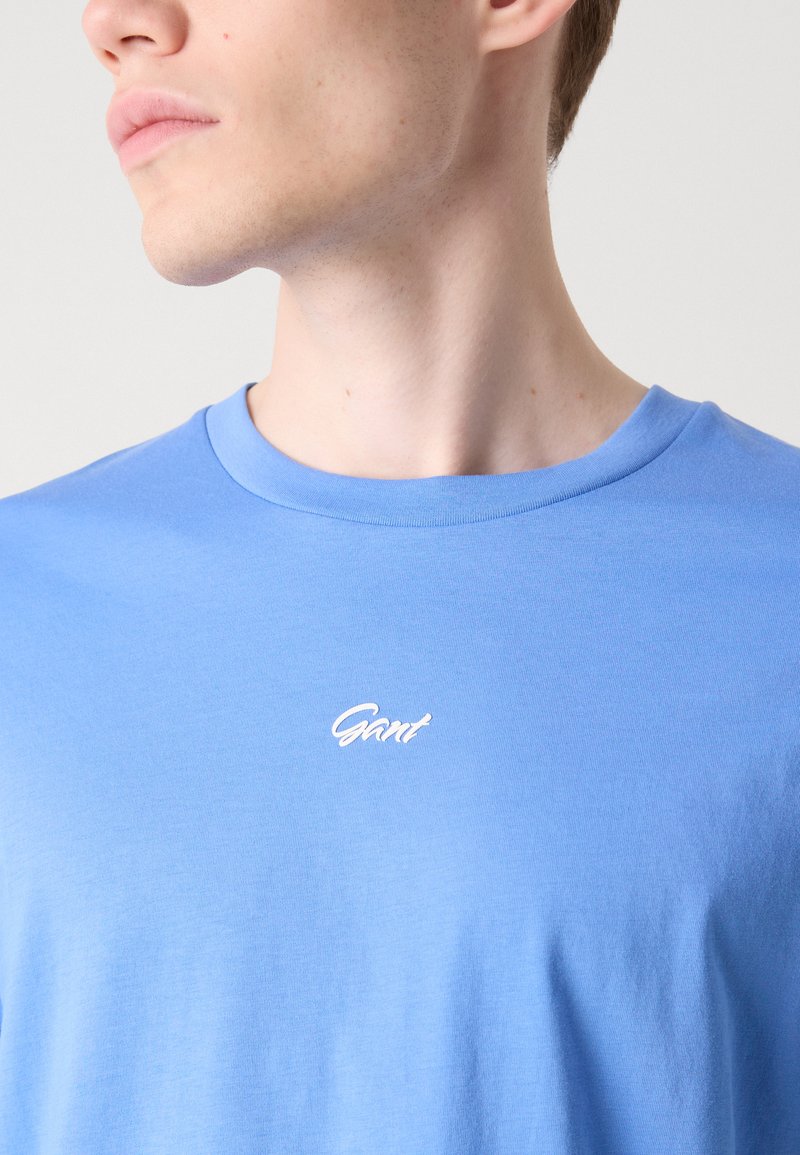 Jasnoniebieska bawełniana koszulka z okrągłym dekoltem, z małym białym logo haftowanym na piersi. Miękka tekstura i luźny krój.