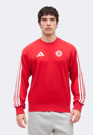 Άνδρας που φοράει κόκκινο φούτερ FC Bayern Munich με λευκές ρίγες Adidas στα μανίκια και γκρι φόρμα, στέκεται μπροστά σε μονόχρωμο φόντο.