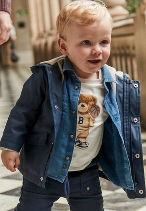 Bambino con capelli biondi che indossa una giacca blu navy e una maglietta con un grafico di orso mentre cammina all'interno su un pavimento a scacchi.