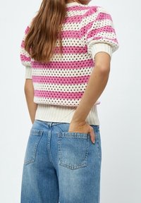 Žena s dlouhými vlasy, oblečená v bílém a růžovém pruhovaném krátkém svetr s krátkým rukávem a modrých denimových džínech, má ruku v kapsičce na zadku.