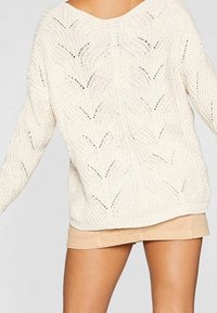 Pull crème en tricot épais avec un col en V, présentant un motif de mailles ajourées et un ourlet côtelé. Associé à une mini jupe beige.