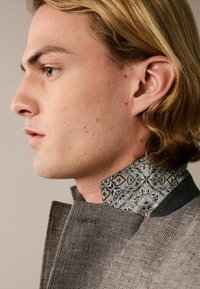 Braunes, strukturiertes Kleidungsstück mit hohem Kragen; gemusterter Stoff in Blau und Beige am Kragen sichtbar. Subtile Details am Rand der Jacke.