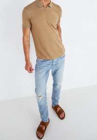 En tanfärgad pikétröja med dragkedja, ljusblå ripped jeans och bruna slidesandaler. Outfitten har en smal passform och avslappnad stil.