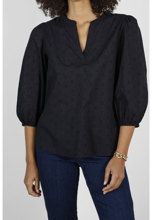 Woman wearing a black blouse with subtle textured pattern and 3/4 puff sleeves paired with dark blue jeans and a gold chain bracelet.