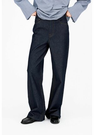 Personne portant un jean large foncé, des chaussures noires et une chemise à manches longues bleue à carreaux, les mains dans les poches arrière.