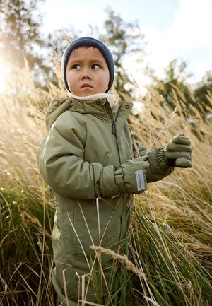 Manteau matelassé vert avec zip et capuche, col en tricot blanc, portant un bonnet bleu et des gants verts, debout dans de l'herbe haute.