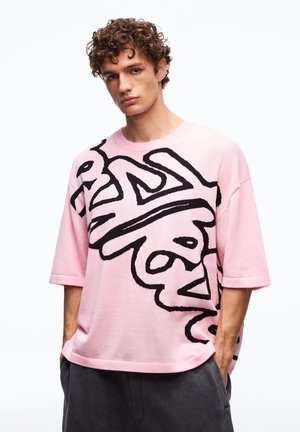 Młody mężczyzna z kręconymi włosami nosi luźną różową koszulę z dużymi czarnymi abstrakcyjnymi literami, ręce w kieszeniach, na jednolitym białym tle.