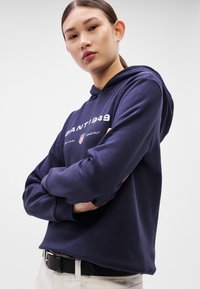 Námořnicky modrá mikina s bílým textovým logem přes hrudník, dlouhými rukávy a přední kapsou ve stylu klokanky, kombinovaná se světlými kalhotami a černým pásem.