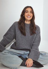 Pull en maille torsadée gris avec un col montant, coupe décontractée et manches longues, associé à un jean large bleu clair et des talons noirs.