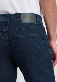 Donkerblauwe spijkerbroek met een achterzak voorzien van een subtiel V-stikselpatroon. Leren label op de tailleband met "pierre cardin" branding.