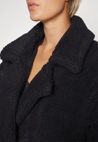 Cappotto di pile nero con una superficie texturizzata e morbida, caratterizzato da un ampio colletto e una vista ravvicinata del scollo.