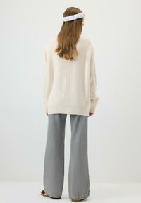 Pikkade juustega inimene, kes kannab valget kootud kampsunit, laiu halli püksid ja valget punutud peapaela, seisab tavalise tausta ees.