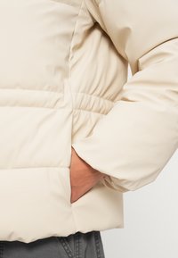 Beige pufferjakke med polstret design, elastisk talje og sidelomme. Glat, let skinnende tekstil med detaljeret syning.