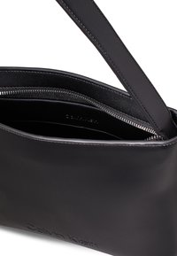 Borsa in pelle nera con superficie liscia, chiusura con cerniera e una sola tracolla. Presenta un logo impresso all'esterno. L'interno è foderato in tessuto nero.