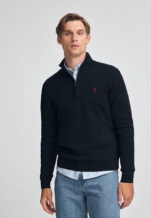 Sweatshirt noir zippé en matériau tricoté, présentant un poignet et un ourlet côtelés. Il possède un petit logo rouge sur la poitrine gauche. Porté sur une chemise bleu clair.