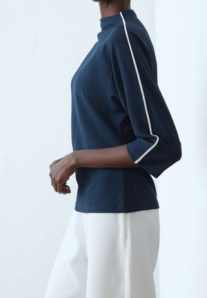 Personne portant un haut à manches longues bleu marine avec une bande blanche le long de la manche et un pantalon large blanc, debout de profil sur un fond uni.