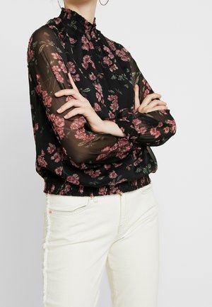 Personne portant un chemisier noir transparent à motifs floraux avec manches longues et pantalon blanc, bras croisés, sur un fond clair uni.