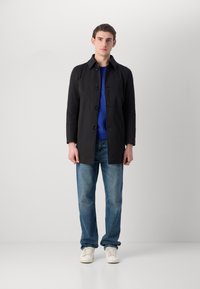 Cappotto nero su misura con taglio dritto, dotato di chiusura a bottoni, colletto appuntito e due tasche frontali, indossato sopra una camicia blu e jeans.