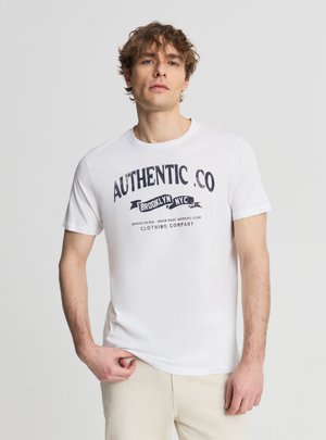 Giovane con capelli ricci che indossa una maglietta bianca con testo nero "AUTHENTIC CO Brooklyn NYC", in piedi davanti a uno sfondo chiaro semplice.