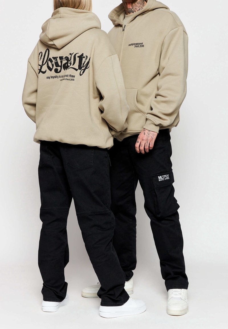 Beige mikina s kapucí a černým grafickým textem na zádech. Černé cargo kalhoty s bočními kapsami a logem. Bílá obuv dotváří celý outfit.