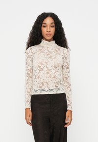 Top in pizzo a maniche lunghe di colore crema, con collo alto e motivo floreale, abbinato a pantaloni neri con motivo.