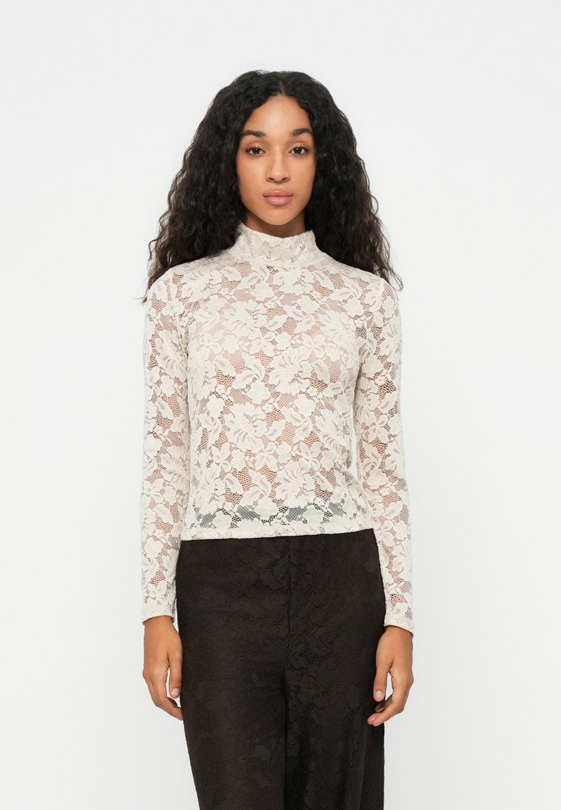 Top in pizzo a maniche lunghe di colore crema, con collo alto e motivo floreale, abbinato a pantaloni neri con motivo.
