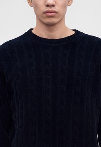 Homme portant un pull en maille torsadée bleu marine foncé avec un col rond sur fond gris clair.