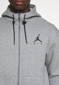 Szara bluza z kapturem wykonana z miękkiego materiału, z zamkiem błyskawicznym z przodu, czarnymi sznurkami do ściągacza oraz czarnym haftowanym logo Jumpman na piersi.