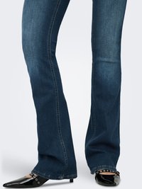 Donkerblauwe jeans met uitlopende pijpen en een gladde textuur, geaccentueerd met zichtbare stiksels. Gecombineerd met glanzende zwarte schoenen met puntige neus en decoratieve hardware.