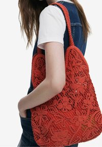 Rød hæklet taske med blomster mønstre, åbent vævet design og en robust flettet rem, båret under armen.