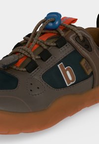 Baskets marron et noires avec des accents orange, des lacets élastiques maintenus par un verrou bleu, et une semelle couleur gomme.